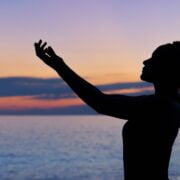 A silhouette of a woman with her hands raised in the air at sunset.