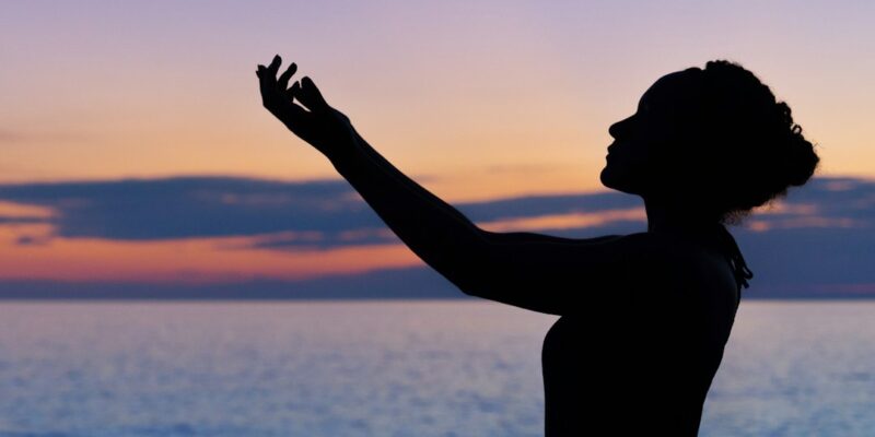 A silhouette of a woman with her hands raised in the air at sunset.