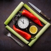 A clock, peppers, lemon, knife and fork on a dark background.