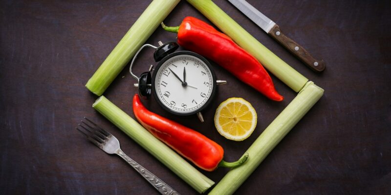 A clock, peppers, lemon, knife and fork on a dark background.