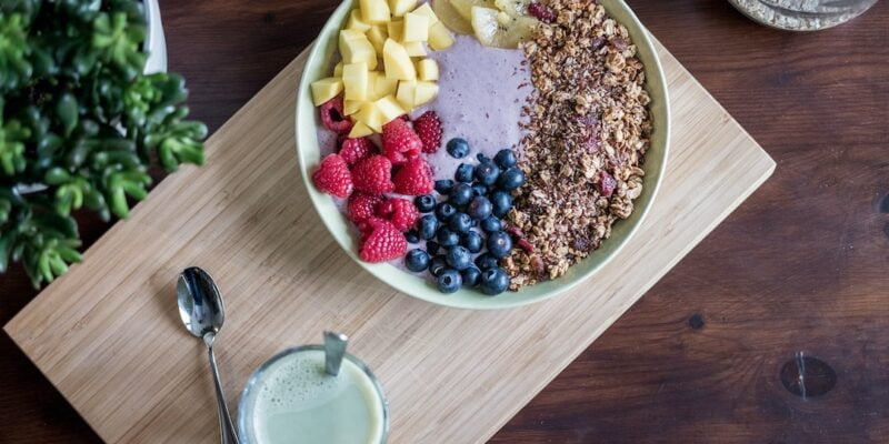 A bowl of fruit and granola on a wooden cutting board.