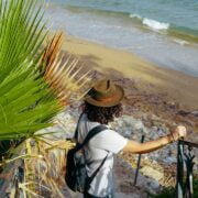 A man wearing a hat is standing on a railing near a beach.