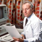 A man in a white lab coat reading a paper.