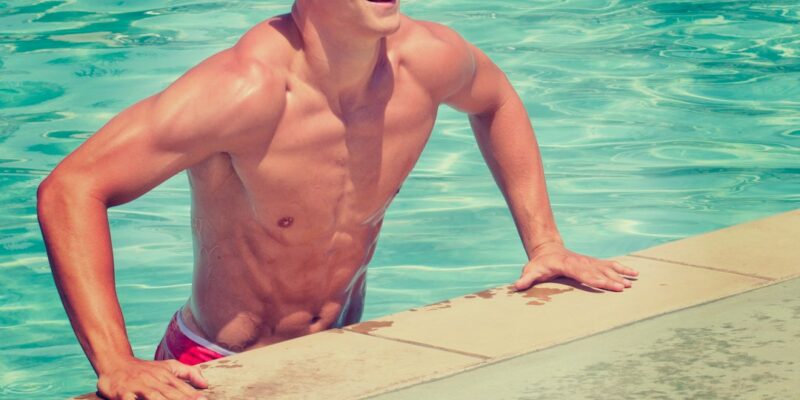 A man in swim trunks leaning on the edge of a pool.