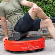 A person in a green shirt and black shorts is sitting on a red vibrating exercise platform labeled "Power Plate MOVE." They are using a black foam roller under their left thigh. The setting includes a stone wall and greenery in the background.