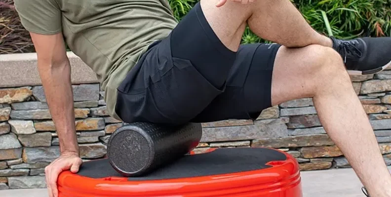 A person in a green shirt and black shorts is sitting on a red vibrating exercise platform labeled "Power Plate MOVE." They are using a black foam roller under their left thigh. The setting includes a stone wall and greenery in the background.