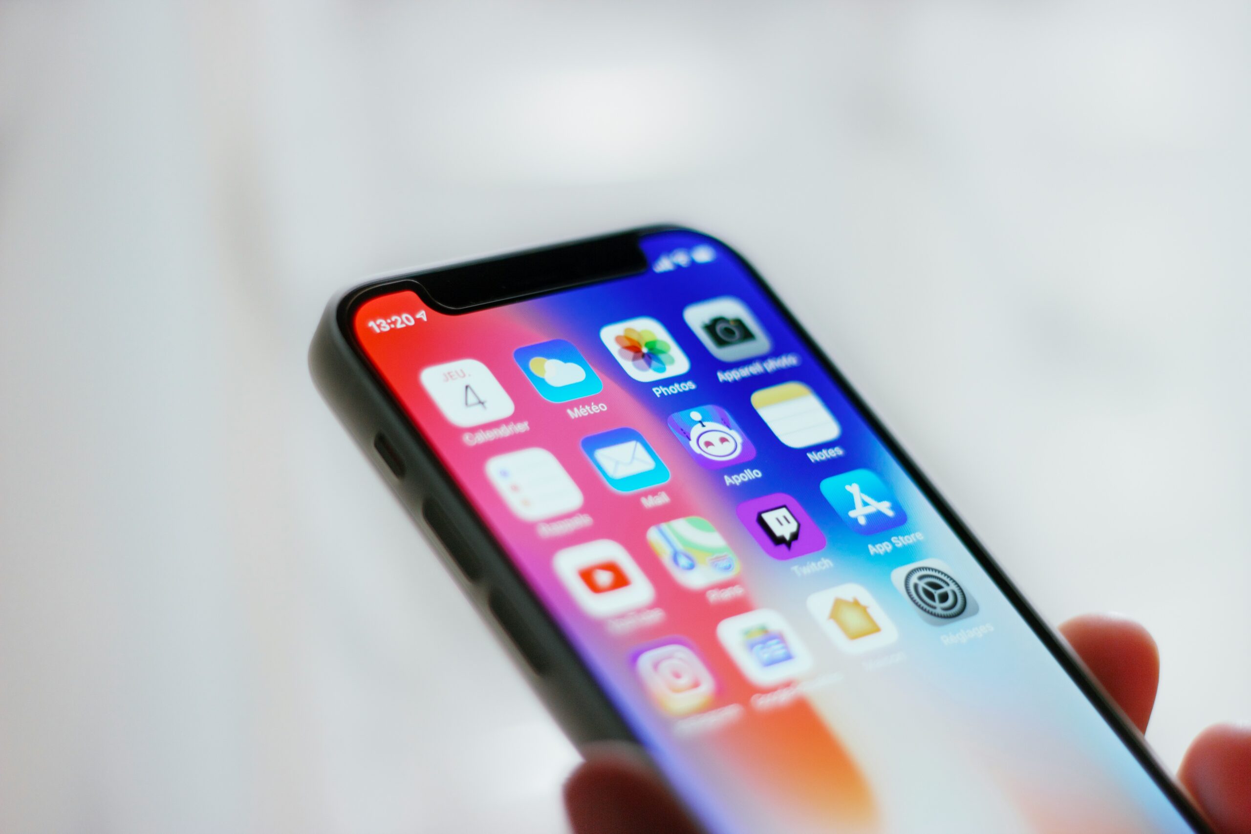 A person holds a smartphone with a colorful display, showing several app icons including Mail, Photos, and App Store. The time reads 13:29, and the background is blurred white, emphasizing the focus on the phone screen. The device has a black edge and slim profile.