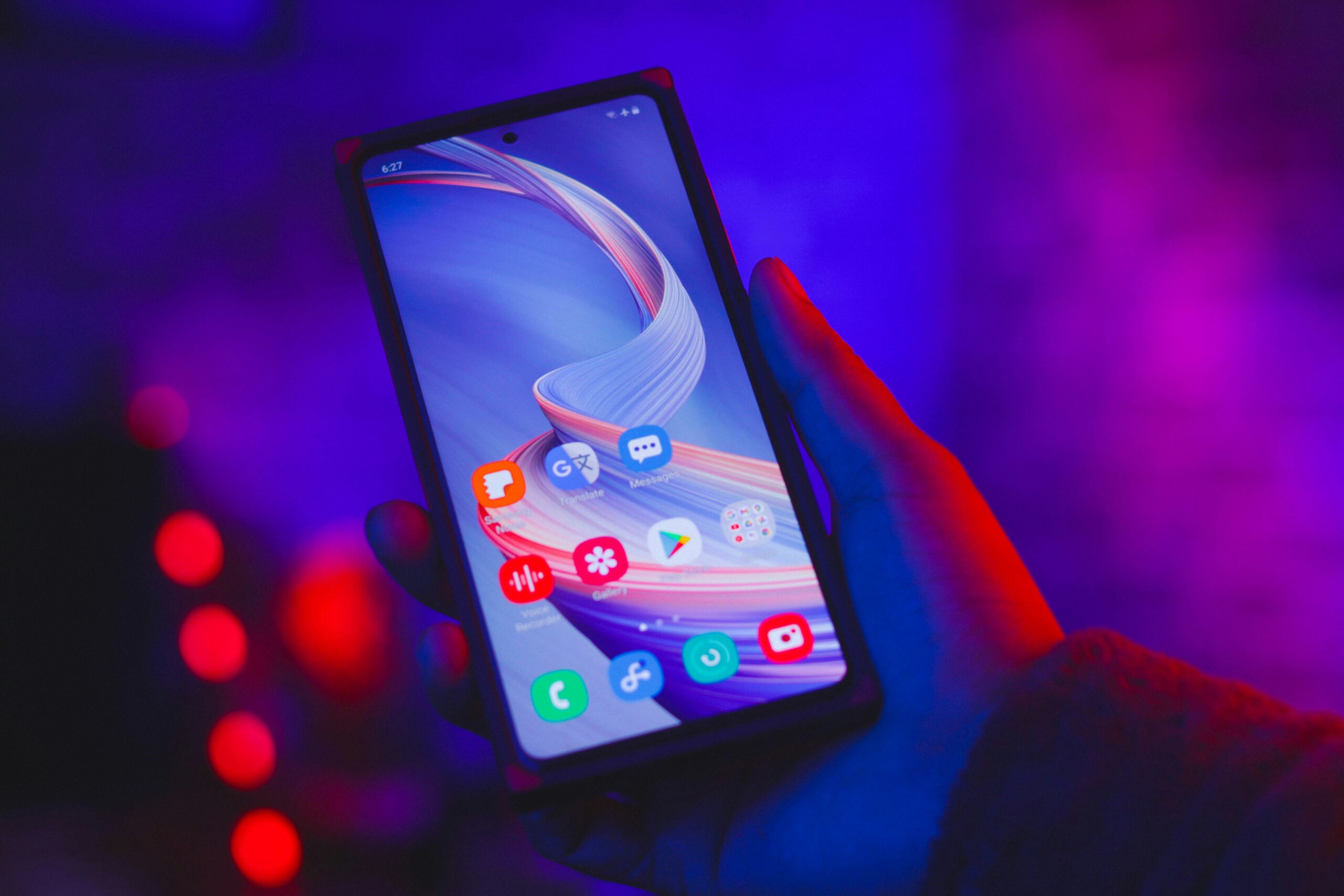 A hand holding a smartphone in a dimly lit setting with a vibrant red and blue background. The phone screen displays various app icons, including social media, messaging, and settings, set against an abstract swirl wallpaper. The time reads 1:07 in the top left corner.