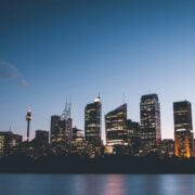 City skyline at dusk with illuminated buildings reflecting over water.