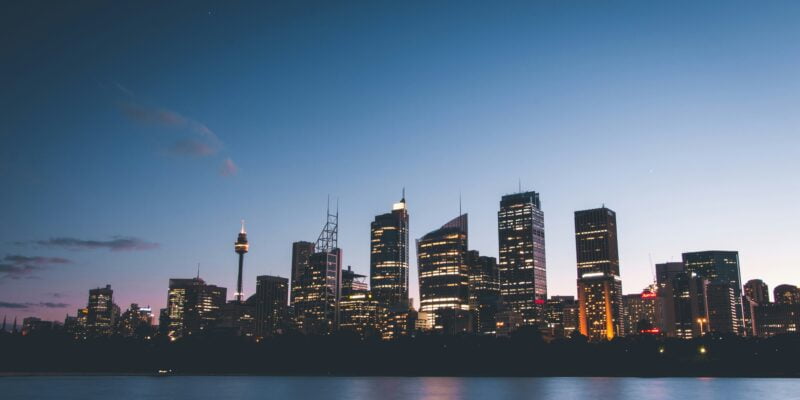 City skyline at dusk with illuminated buildings reflecting over water.