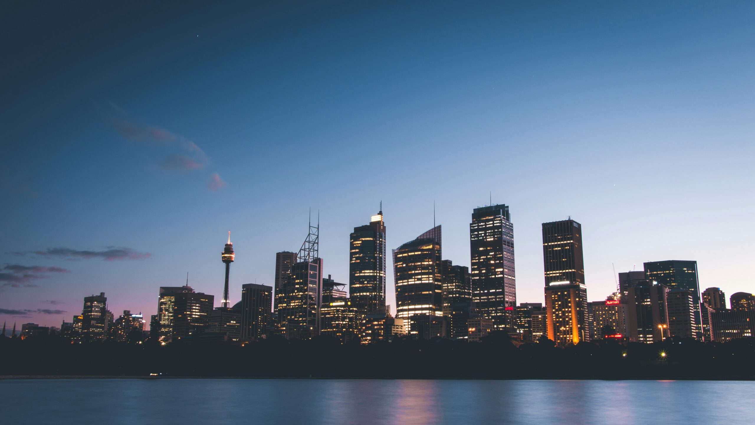 City skyline at dusk with illuminated buildings reflecting over water.