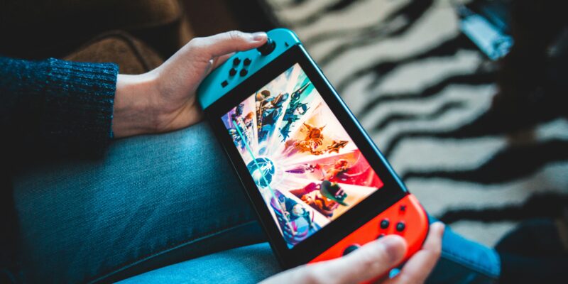 A person holds a Nintendo Switch with blue and red Joy-Con controllers, displaying a colorful game on the screen. The person is seated, wearing jeans and a dark sweater. The background features a sofa and a zebra-striped rug.