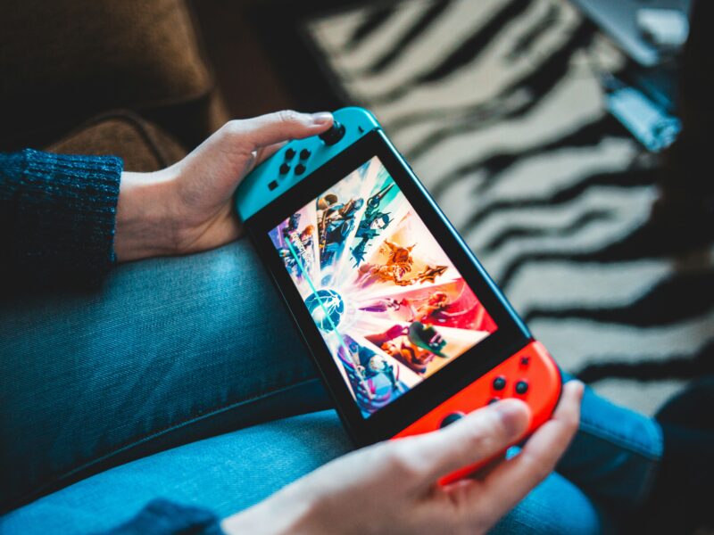 A person holds a Nintendo Switch with blue and red Joy-Con controllers, displaying a colorful game on the screen. The person is seated, wearing jeans and a dark sweater. The background features a sofa and a zebra-striped rug.