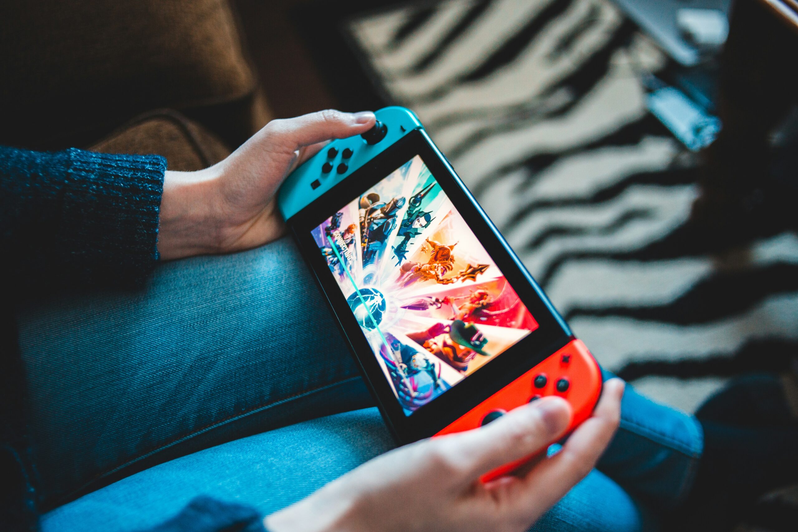 A person holds a Nintendo Switch with blue and red Joy-Con controllers, displaying a colorful game on the screen. The person is seated, wearing jeans and a dark sweater. The background features a sofa and a zebra-striped rug.