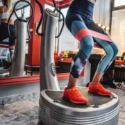 A person is exercising on a vibration plate, wearing colorful leggings and bright orange sneakers. A resistance band is around their thighs. The room has red walls, a large mirror, and several pairs of shoes on display. A window allows natural light to illuminate the space.