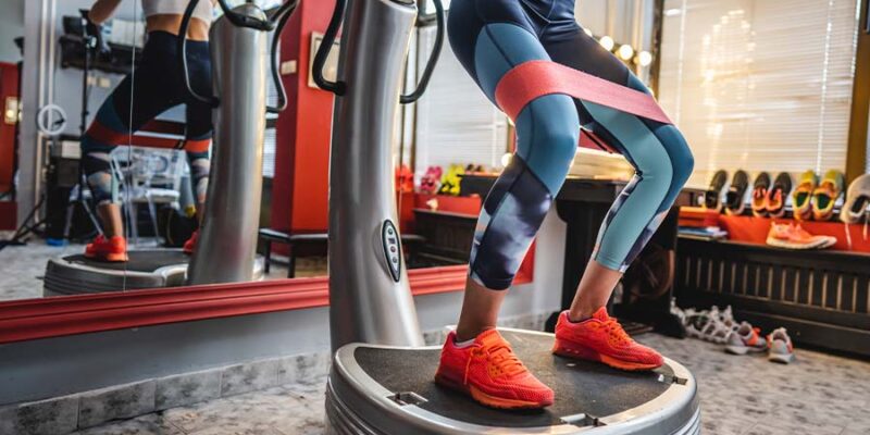 A person is exercising on a vibration plate, wearing colorful leggings and bright orange sneakers. A resistance band is around their thighs. The room has red walls, a large mirror, and several pairs of shoes on display. A window allows natural light to illuminate the space.