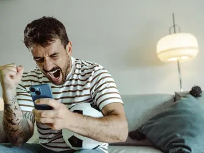 Man excitedly cheering while holding a smartphone and a game controller.