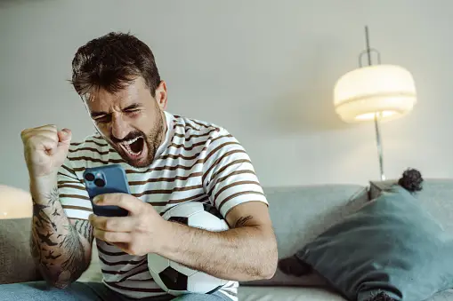 Man excitedly cheering while holding a smartphone and a game controller.