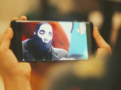 Person holding smartphone while recording someone wearing a mask and suit.