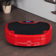 A red Power Plate Move, a vibration exercise machine, is placed on a wooden floor near a potted plant and stairs. The device features a black platform and a digital display showing "00.00". The branding is visible on its side.