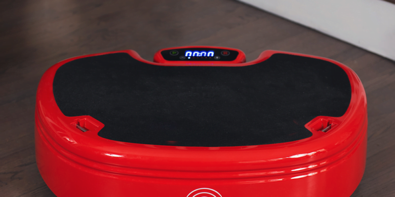 A red Power Plate Move, a vibration exercise machine, is placed on a wooden floor near a potted plant and stairs. The device features a black platform and a digital display showing "00.00". The branding is visible on its side.