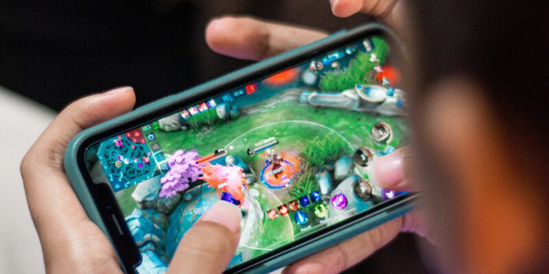 A person wearing a lavender shirt is playing a mobile game on a smartphone. The screen shows a colorful game with various characters and vibrant landscapes. The person is using both thumbs to control the game, focusing intently on the screen.