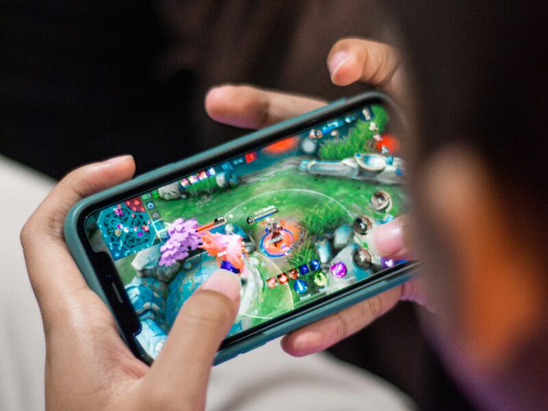 A person wearing a lavender shirt is playing a mobile game on a smartphone. The screen shows a colorful game with various characters and vibrant landscapes. The person is using both thumbs to control the game, focusing intently on the screen.