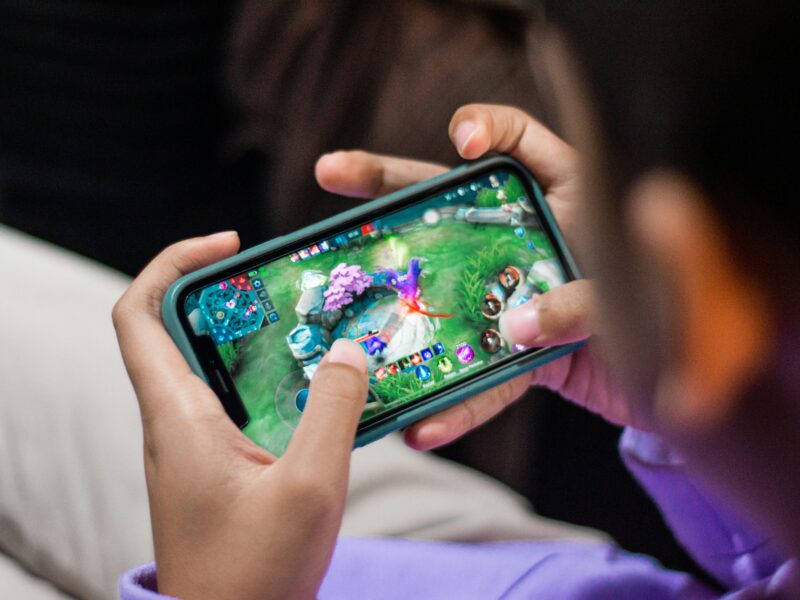 A person holds a smartphone horizontally, playing a colorful video game featuring characters and landscapes on the screen. The game includes health bars and a map in the top corners. The person's hands and part of a purple sleeve are visible, along with a blurred background.