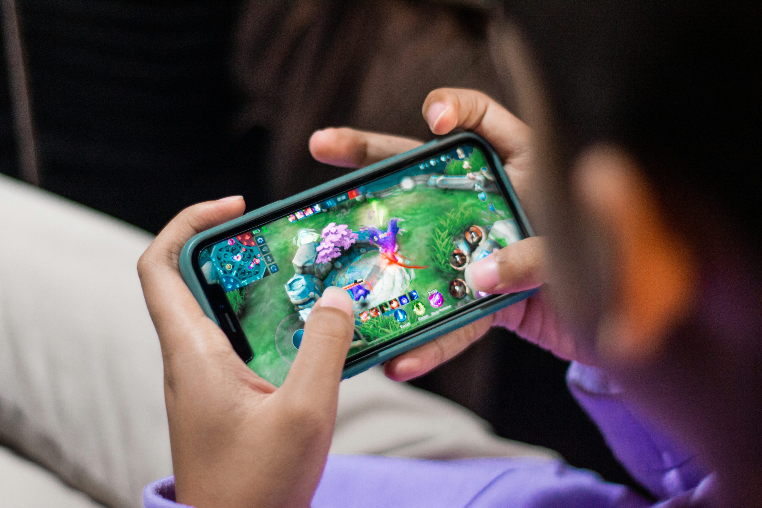 A person holds a smartphone horizontally, playing a colorful video game featuring characters and landscapes on the screen. The game includes health bars and a map in the top corners. The person's hands and part of a purple sleeve are visible, along with a blurred background.