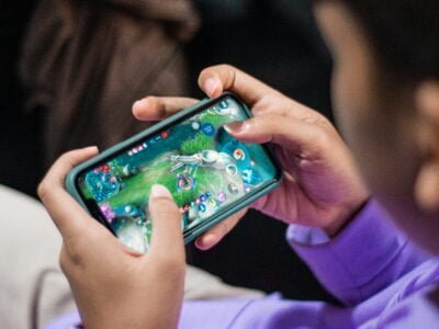 A person wearing a purple shirt is holding a smartphone horizontally, playing a video game. The screen displays a colorful, action-packed game interface with various icons and controls. The person's thumbs are actively tapping the screen, suggesting engagement in the gameplay.