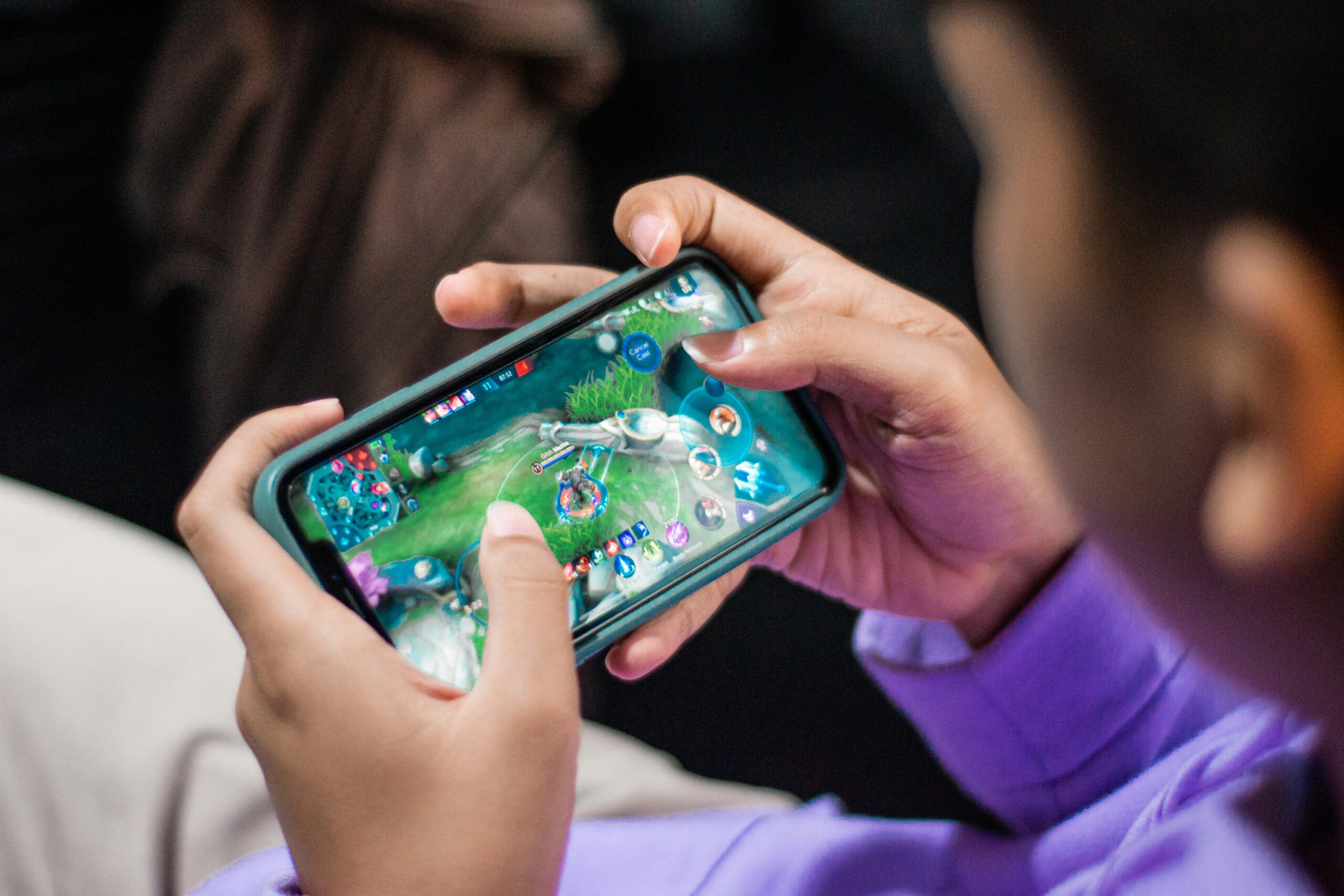 A person wearing a purple shirt is holding a smartphone horizontally, playing a video game. The screen displays a colorful, action-packed game interface with various icons and controls. The person's thumbs are actively tapping the screen, suggesting engagement in the gameplay.