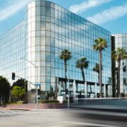 A modern glass-fronted building with palm trees reflected in its facade, viewed from across a street with blurred traffic in the foreground.