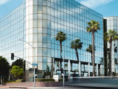 A modern glass-fronted building with palm trees reflected in its facade, viewed from across a street with blurred traffic in the foreground.