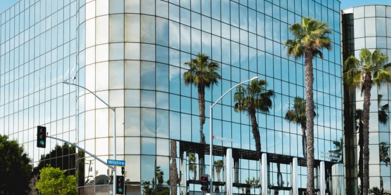 A modern glass-fronted building with palm trees reflected in its facade, viewed from across a street with blurred traffic in the foreground.