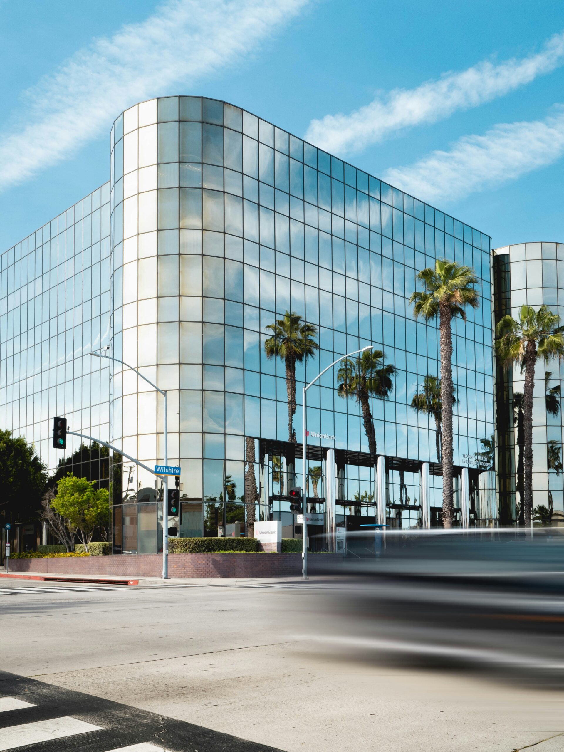 A modern glass-fronted building with palm trees reflected in its facade, viewed from across a street with blurred traffic in the foreground.