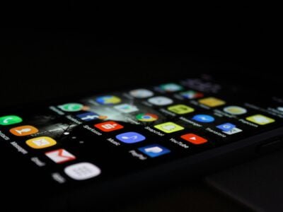 Close-up of a smartphone screen displaying various app icons in a dark environment. Icons include email, music, social media, and navigation apps, arranged in a grid. The screen is glowing brightly, highlighting the colorful icons against the black background.