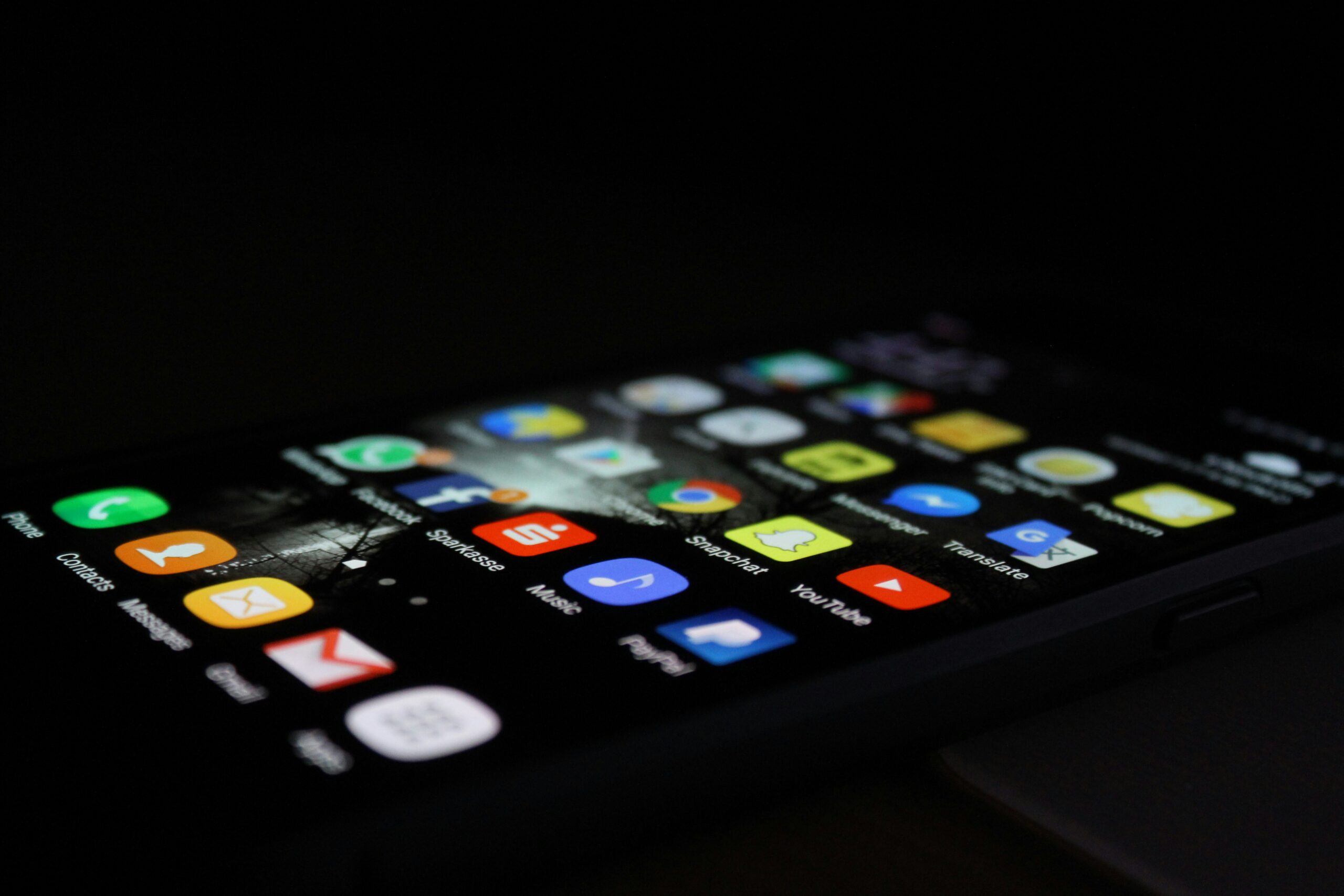 Close-up of a smartphone screen displaying various app icons in a dark environment. Icons include email, music, social media, and navigation apps, arranged in a grid. The screen is glowing brightly, highlighting the colorful icons against the black background.