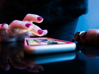 Close-up of a hand with sparkly purple nail polish using a smartphone on a reflective black table. The smartphone's screen displays colorful app icons. A brown leather bag is partially visible on the right. The lighting gives the scene a cool-toned ambiance.