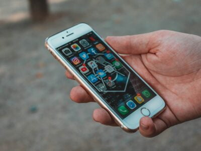 A hand holds a white smartphone displaying various apps on the home screen, including Facebook, Instagram, Twitter, and WhatsApp. The phone has a cracked glass screen. The background is a blurred outdoor setting with a brownish ground surface.