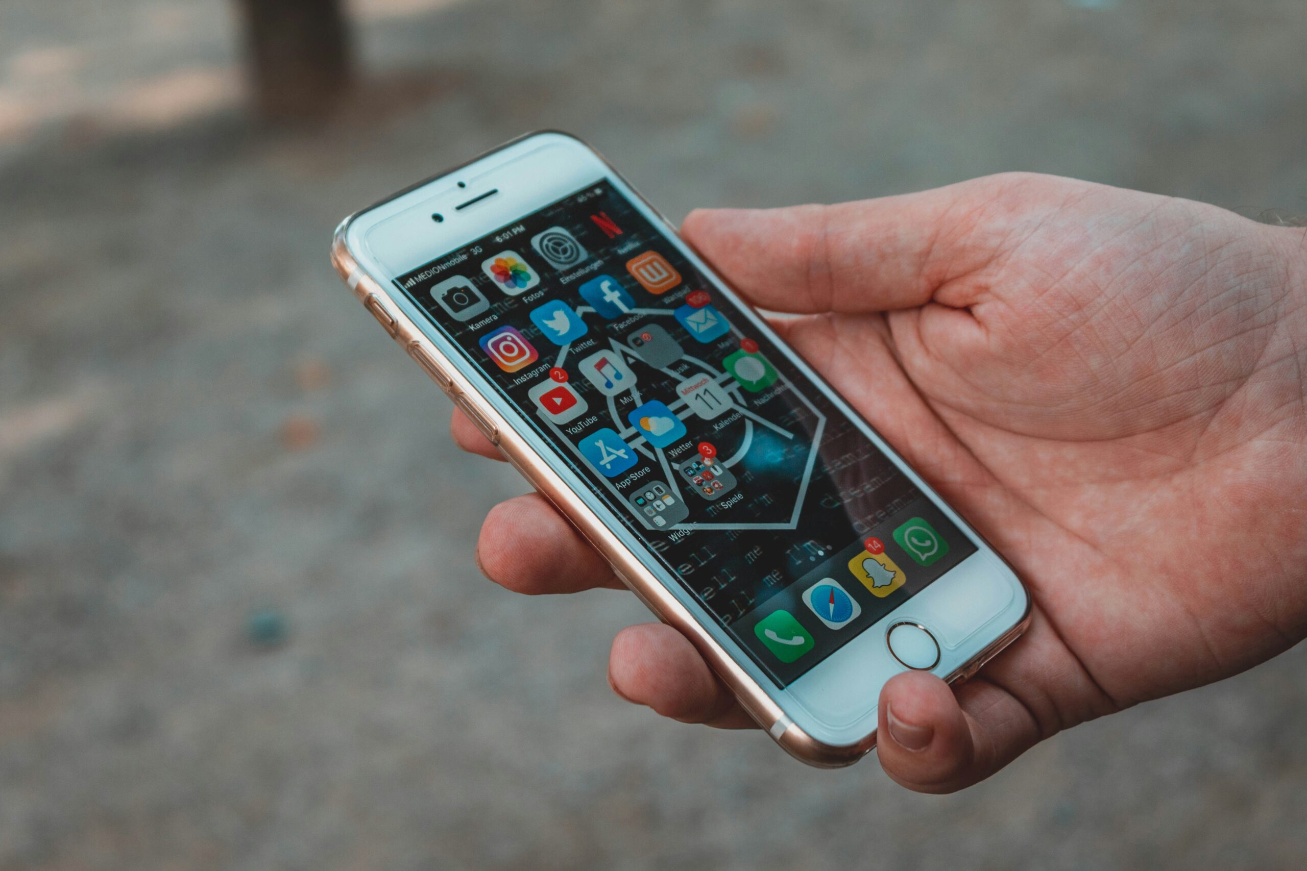 A hand holds a white smartphone displaying various apps on the home screen, including Facebook, Instagram, Twitter, and WhatsApp. The phone has a cracked glass screen. The background is a blurred outdoor setting with a brownish ground surface.