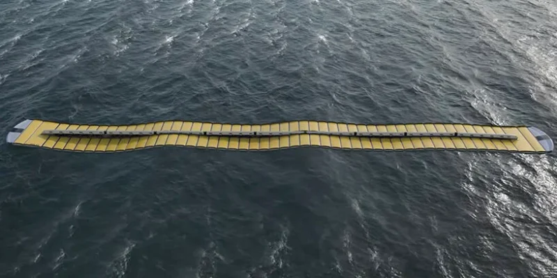 An aerial view shows a long, yellow wave energy converter floating on the ocean. This device, resembling wave magnets with its segmented, undulating structure, harnesses ocean power from choppy seas and white-capped waves, promising a sustainable future with each surge it captures.