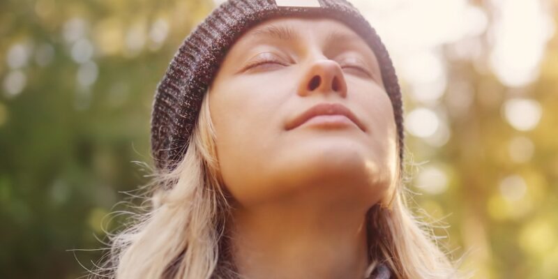 A woman in a beanie looking up at the sun in the woods, contemplating why Facebook is down.