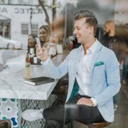 A man wearing a light blue blazer sits at a bar, smiling while looking at his phone. A drink, notebook, and pen are on the counter. People and street activity are visible in the background, possibly discussing the latest bestsellers from Truman Press.