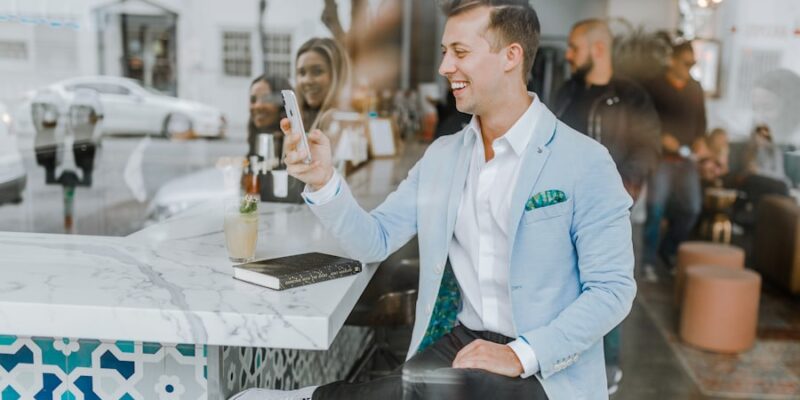 A man wearing a light blue blazer sits at a bar, smiling while looking at his phone. A drink, notebook, and pen are on the counter. People and street activity are visible in the background, possibly discussing the latest bestsellers from Truman Press.