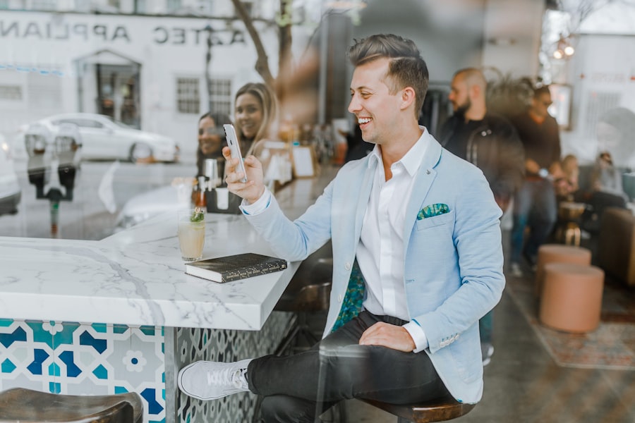 A man wearing a light blue blazer sits at a bar, smiling while looking at his phone. A drink, notebook, and pen are on the counter. People and street activity are visible in the background, possibly discussing the latest bestsellers from Truman Press.