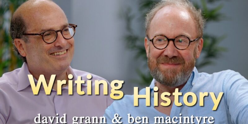 Two men, each wearing glasses, sit and smile in a room with the text "Writing History" and "david grann & ben macintyre" written on the image.