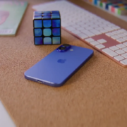 A blue smartphone, a Rubik's Cube, and a wireless keyboard are placed on a cork surface, along with a partially visible magazine.