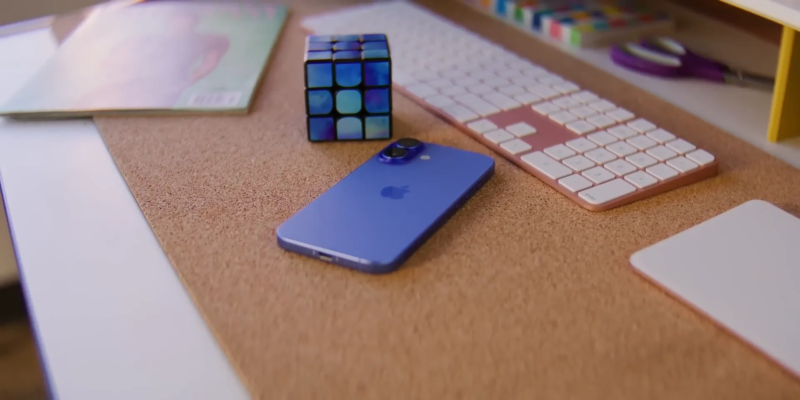 A blue smartphone, a Rubik's Cube, and a wireless keyboard are placed on a cork surface, along with a partially visible magazine.