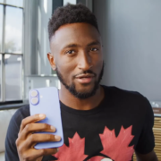 A man in a black shirt with a red maple leaf design holds a blue smartphone, standing in a well-lit modern room.