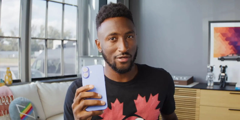 A man in a black shirt with a red maple leaf design holds a blue smartphone, standing in a well-lit modern room.